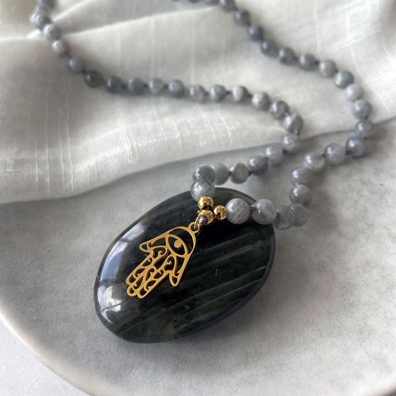 Close-up of a Mala made of Labradorite gemstones and a gold plated Hamsa Hand as a pendant placed on a Labradorite gemstone on a grey ceramic plate.