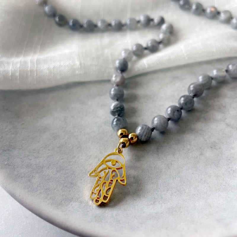 Close-up of a Mala made of Labradorite gemstones and a gold plated Hamsa Hand as a pendant on a grey ceramic plate.