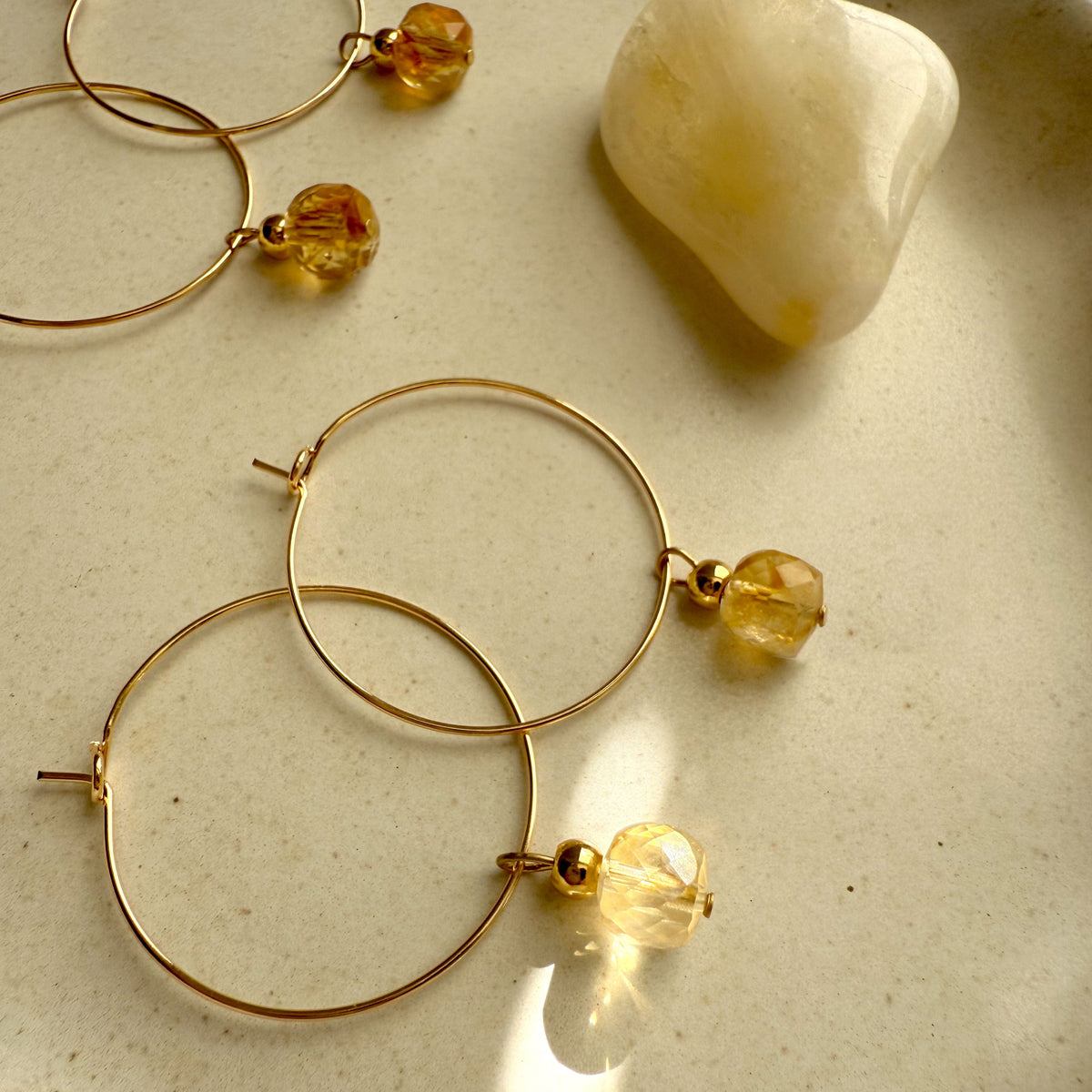 Gold hoop earrings with faceted citrine gemstone charms, glowing in warm sunlight beside a polished citrine crystal on a neutral ceramic dish — handcrafted Halo Hoop earrings by Moksha Jewelry.