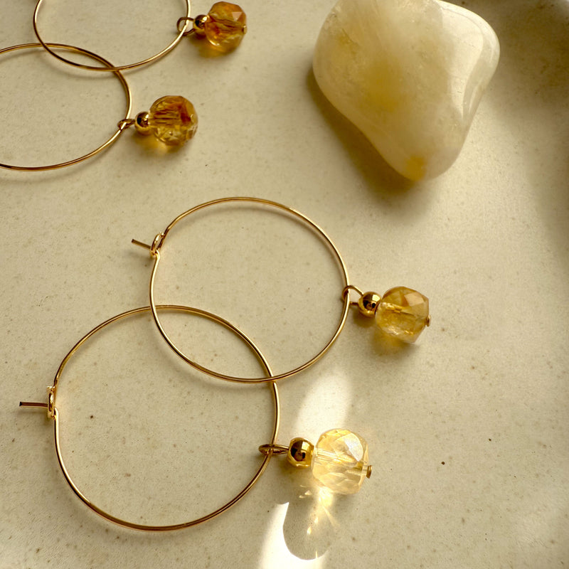 Gold hoop earrings with faceted citrine gemstone charms, glowing in warm sunlight beside a polished citrine crystal on a neutral ceramic dish — handcrafted Halo Hoop earrings by Moksha Jewelry.