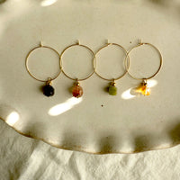 Four pairs of gold hoop earrings with gemstone charms in citrine, green jade, red hematoid, and amethyst, displayed on a neutral ceramic dish under soft natural sunlight — handmade Halo Hoop collection by Moksha Jewelry.