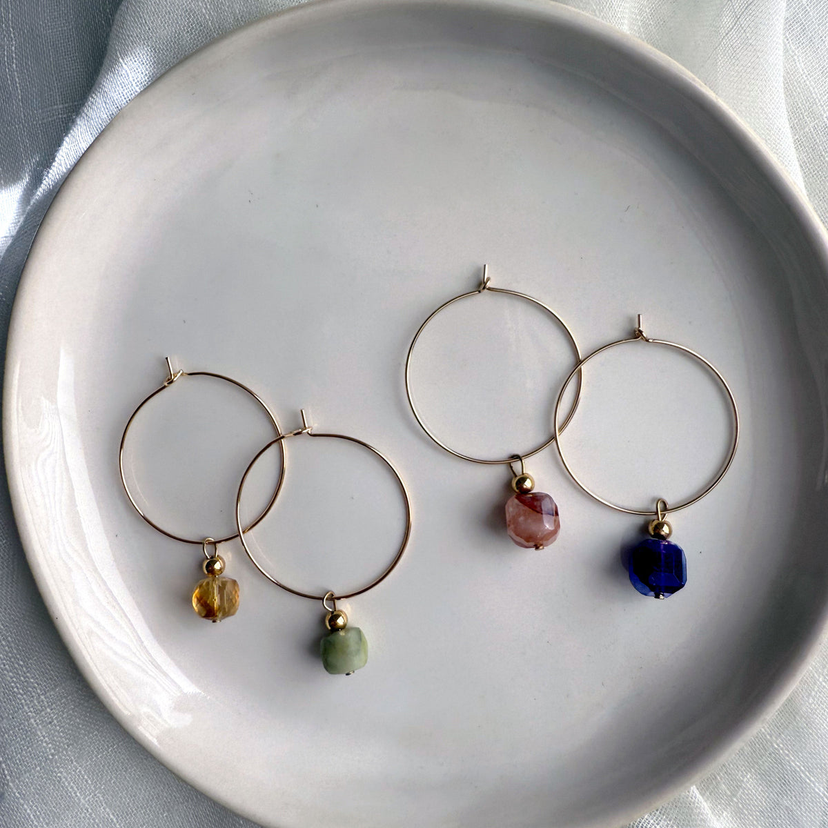 Gold-filled hoop earrings with gemstone charms on a white ceramic plate, featuring citrine, green jade, red hematoid quartz, and amethyst stones from left to right.