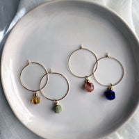 Gold-filled hoop earrings with gemstone charms on a white ceramic plate, featuring citrine, green jade, red hematoid quartz, and amethyst stones from left to right.