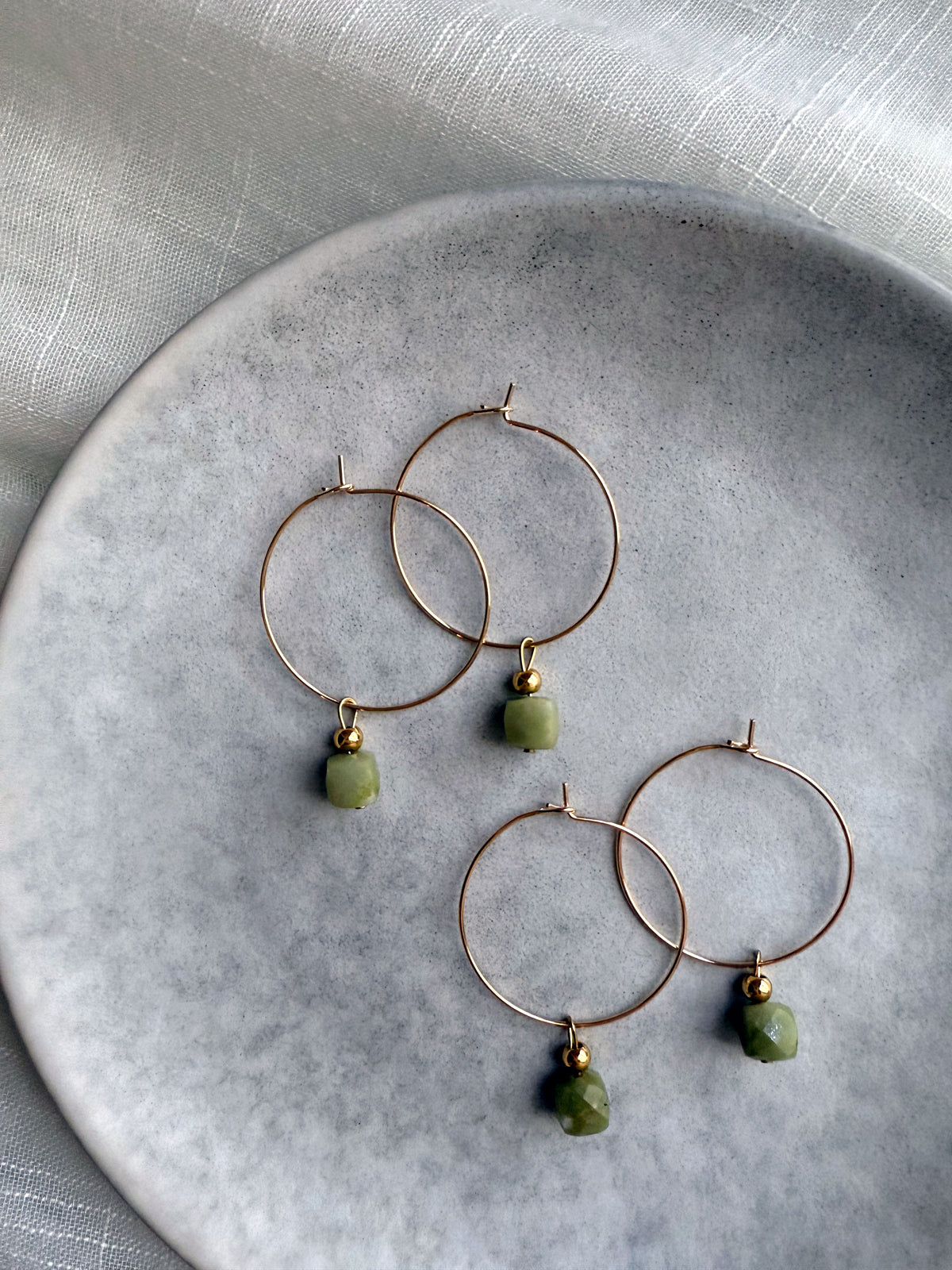 Gold hoop earrings with natural green jade gemstone charms, displayed on a soft grey ceramic plate with elegant fabric background — handcrafted Halo Hoop earrings by Moksha Jewelry.