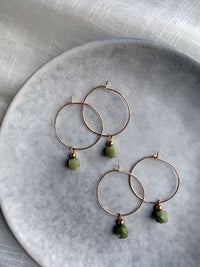 Gold hoop earrings with natural green jade gemstone charms, displayed on a soft grey ceramic plate with elegant fabric background — handcrafted Halo Hoop earrings by Moksha Jewelry.
