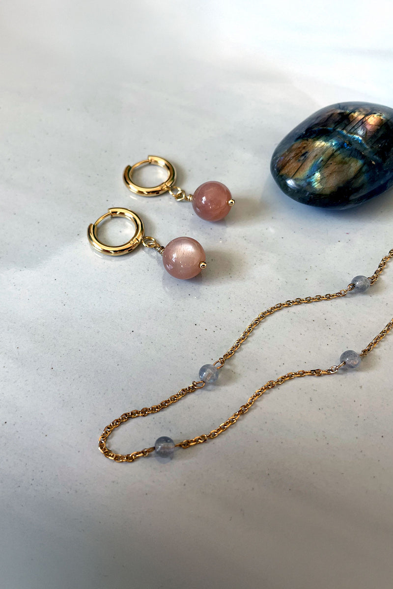 A pair of earrings with a 10mm peach Moonstone and a delicate gold plated chain with 4mm labradorite beads next to a labradorite gemstone on a gray ceramic plate.