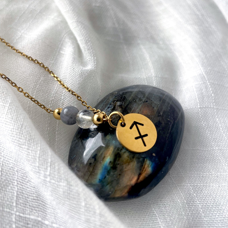 A close-up of a fine golden necklace with a Labradorite and a Citrine bead and a Sagittarius Zodiac pendant laying on a light fabric. The pendant is draped on a Labradorite gemstone.