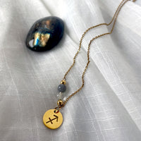 A close-up of a fine golden necklace with a Labradorite and a Citrine bead and a Sagittarius Zodiac pendant laying on a light fabric. Next to the necklace is a labradorite gemstone.