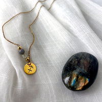 A  fine golden necklace with a Labradorite and a Citrine bead and a Sagittarius Zodiac pendant laying on a light fabric. Next to the necklace is a labradorite gemstone.