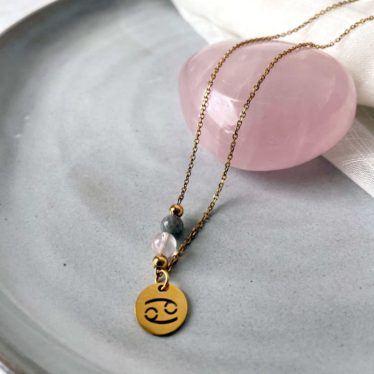 A close-up of a fine necklace with a gold plated Cancer Zodiac charm, a Rose Quartz and a Labradorite bead on a fine gold plated chain, laying on a grey ceramic plate.