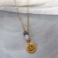 A close-up of a fine necklace with a gold plated Cancer Zodiac charm, a Rose Quartz and a Labradorite bead on a fine gold plated chain, laying on a grey ceramic plate.