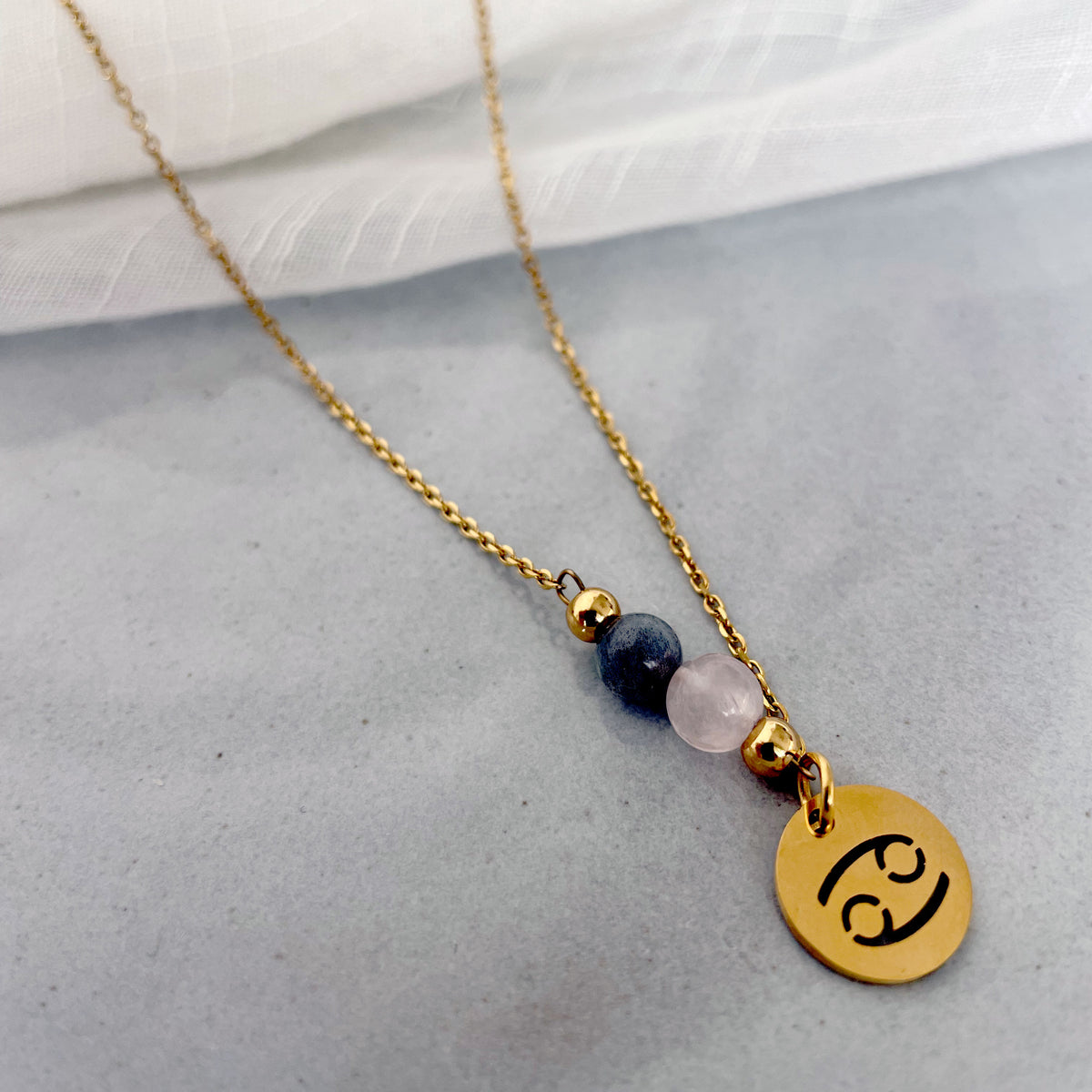 A close-up of a fine necklace with a gold plated Cancer Zodiac charm, a Rose Quartz and a Labradorite bead on a fine gold plated chain, laying on a grey ceramic plate.