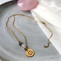 A fine necklace with a gold plated Cancer Zodiac charm, a Rose Quartz and a Labradorite bead on a fine gold plated chain, laying on a grey ceramic plate.