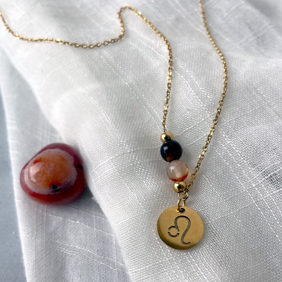 A close-up of a fine golden necklace with a Carnelian and a Tiger Eye bead and a Leo Zodiac pendant laying on a light fabric. Next to the necklace is a Carnelian gemstone.