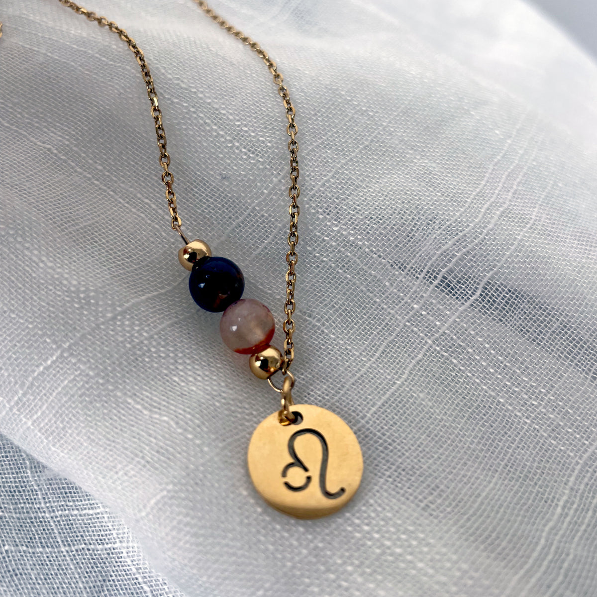 A close-up of a fine golden necklace with a Carnelian and a Tiger Eye bead and a Leo Zodiac pendant laying on a light fabric.