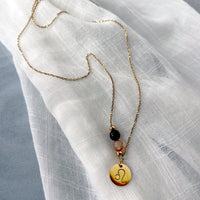 A fine golden necklace with a Carnelian and a Tiger Eye bead and a Leo Zodiac pendant laying on a light fabric.