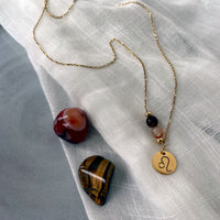 A fine golden necklace with a Carnelian and a Tiger Eye bead and a Leo Zodiac pendant laying on a light fabric. Next to the necklace is a Carnelian and a Tiger Eye gemstone.