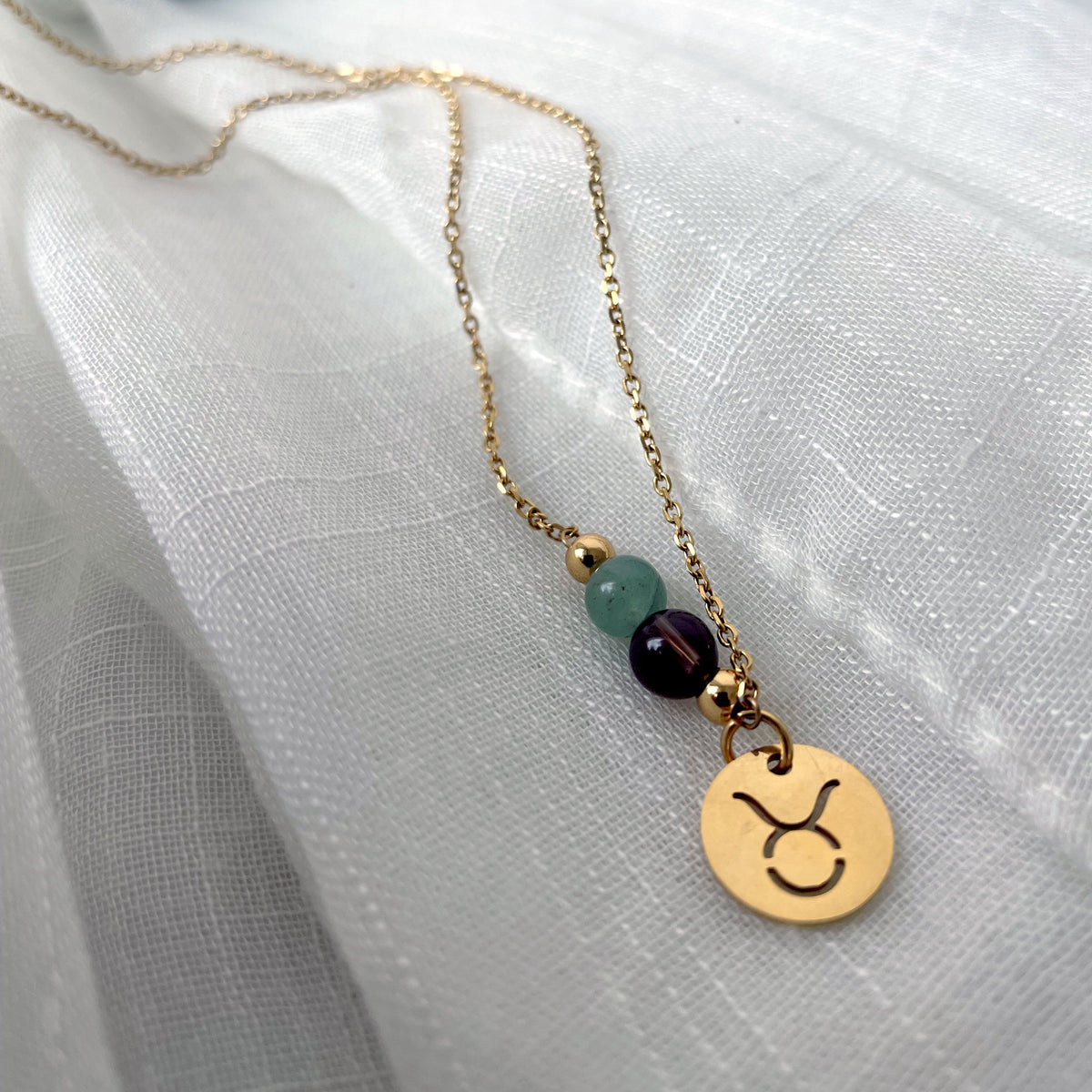 A close-up of a fine golden necklace with a green Aventurine and a Smoky Quartz bead and a Taurus Zodiac pendant laying on a light fabric.