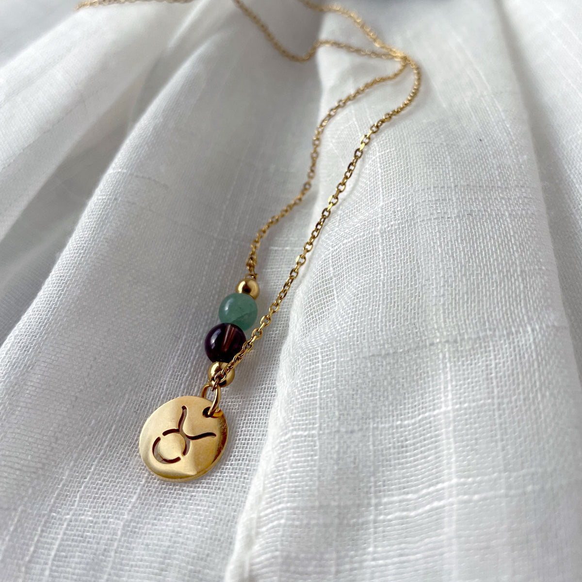 A close-up of a fine golden necklace with a green Aventurine and a Smoky Quartz bead and a Taurus Zodiac pendant laying on a light fabric. 