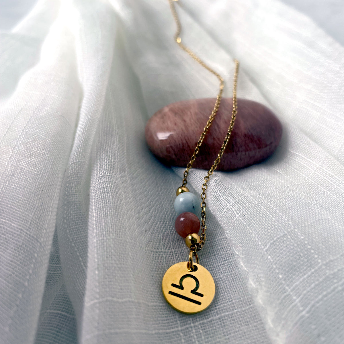 A close-up of a fine golden necklace with a Moonstone and an Aquamarine bead and a Libra Zodiac pendant laying on a light fabric. The necklace is draped on a moonstone gemstone.