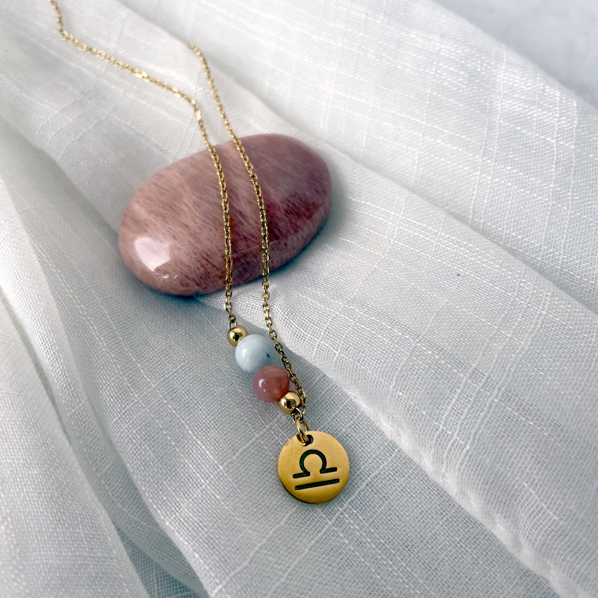 A close-up of a fine golden necklace with a Moonstone and an Aquamarine bead and a Libra Zodiac pendant laying on a light fabric. The necklace is draped on a moonstone gemstone.