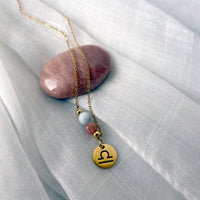 A close-up of a fine golden necklace with a Moonstone and an Aquamarine bead and a Libra Zodiac pendant laying on a light fabric. The necklace is draped on a moonstone gemstone.