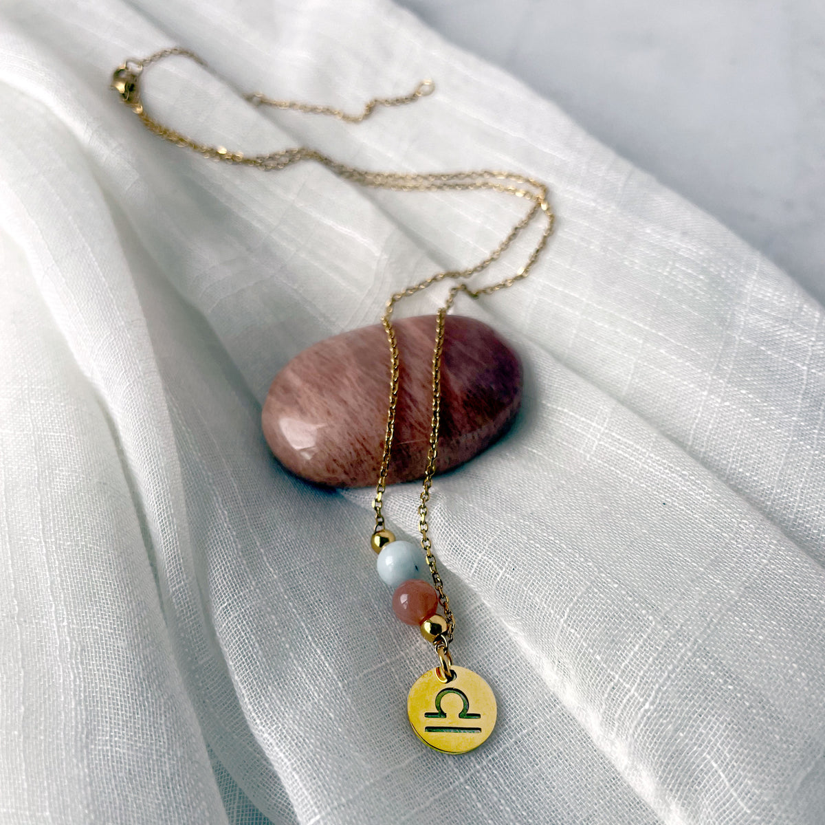 A close-up of a fine golden necklace with a Moonstone and an Aquamarine bead and a Libra Zodiac pendant laying on a light fabric. The necklace is draped on a moonstone gemstone.