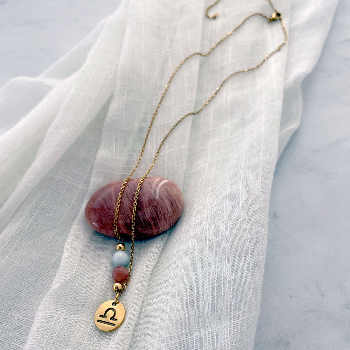 A fine golden necklace with a Moonstone and an Aquamarine bead and a Libra Zodiac pendant laying on a light fabric. The necklace is draped on a moonstone gemstone.