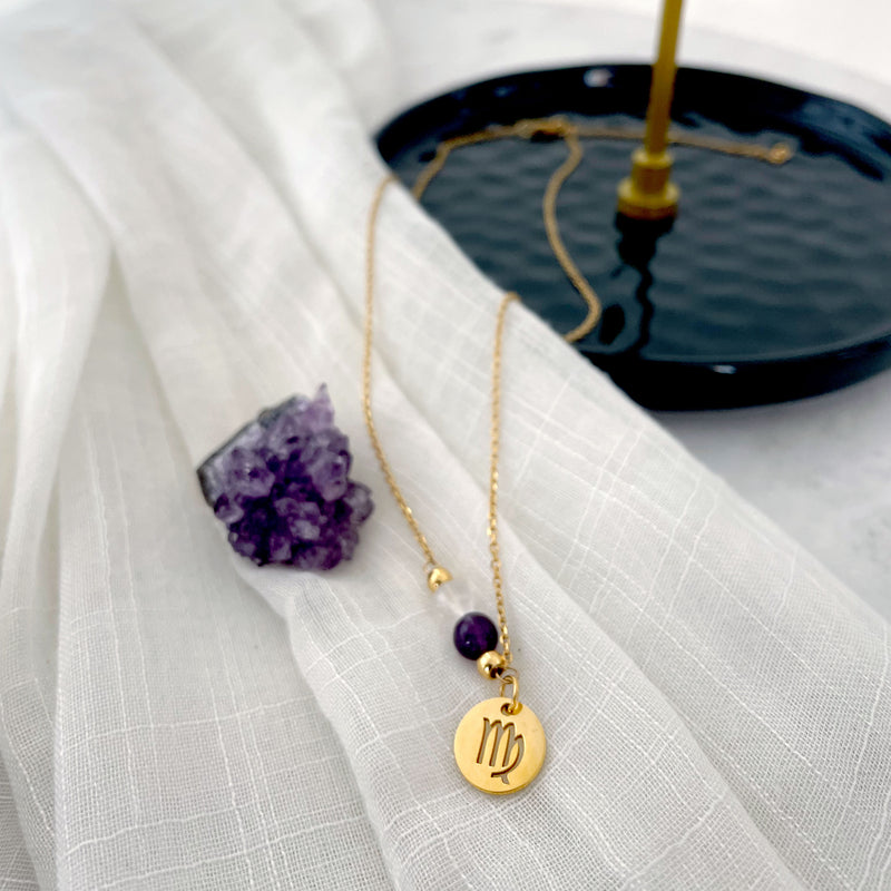 A close-up of a fine golden necklace with a Clear Quartz and an Amethyst bead and a Virgo Zodiac pendant laying on a light fabric. Next to the necklace is an Amethyst druse.