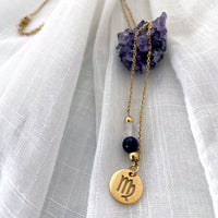 A close-up of a fine golden necklace with a Clear Quartz and an Amethyst bead and a Virgo Zodiac pendant laying on a light fabric. The necklace is draped on an Amethyst druse.