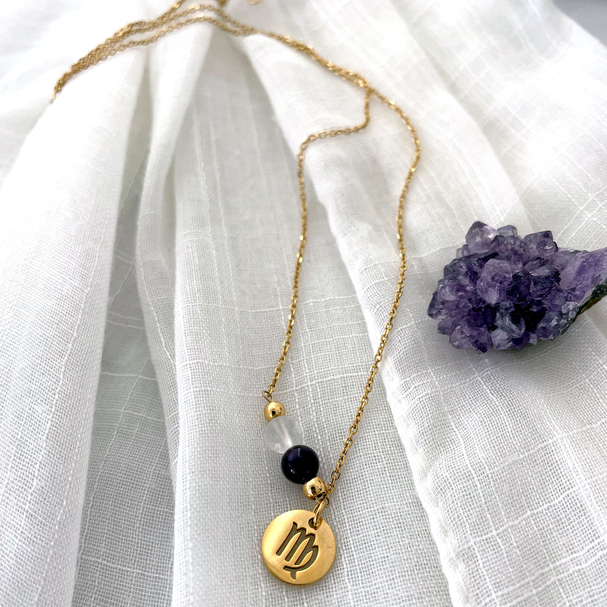 A close-up of a fine golden necklace with a Clear Quartz and an Amethyst bead and a Virgo Zodiac pendant laying on a light fabric. Next to the necklace is an Amethyst druse.