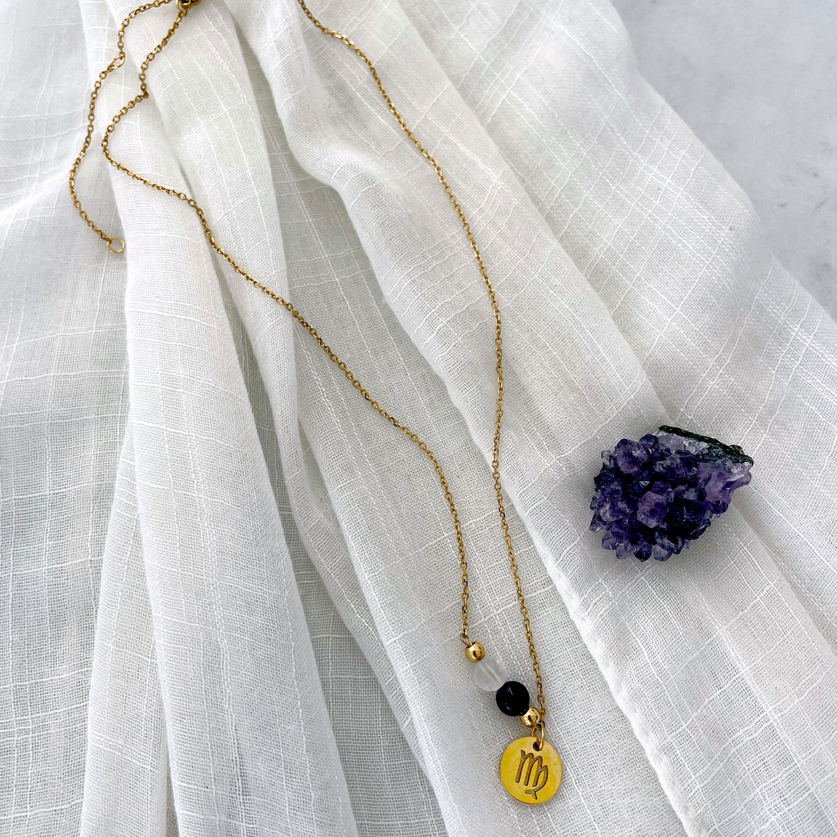 A fine golden necklace with a Clear Quartz and an Amethyst bead and a Virgo Zodiac pendant laying on a light fabric. Next to the necklace is an Amethyst druse.