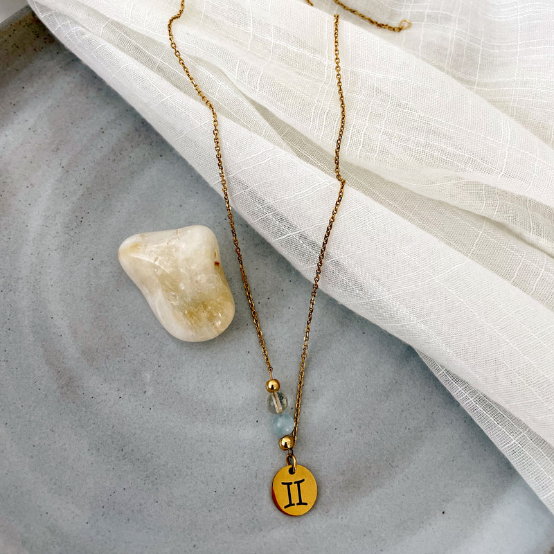 A fine golden necklace with a gemini pendant, a Citrine and a Aquamarine bead on a ceramic plate with a Citrine gemstone.