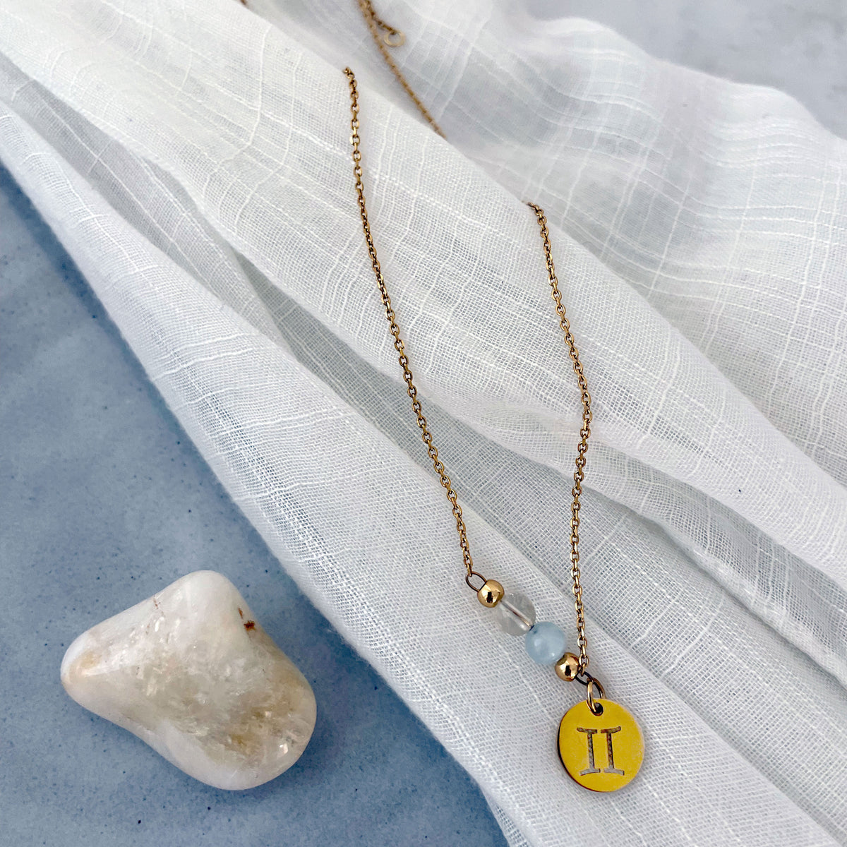 A fine golden necklace with a gemini pendant, a Citrine and a Aquamarine bead on a ceramic plate with a Citrine gemstone.