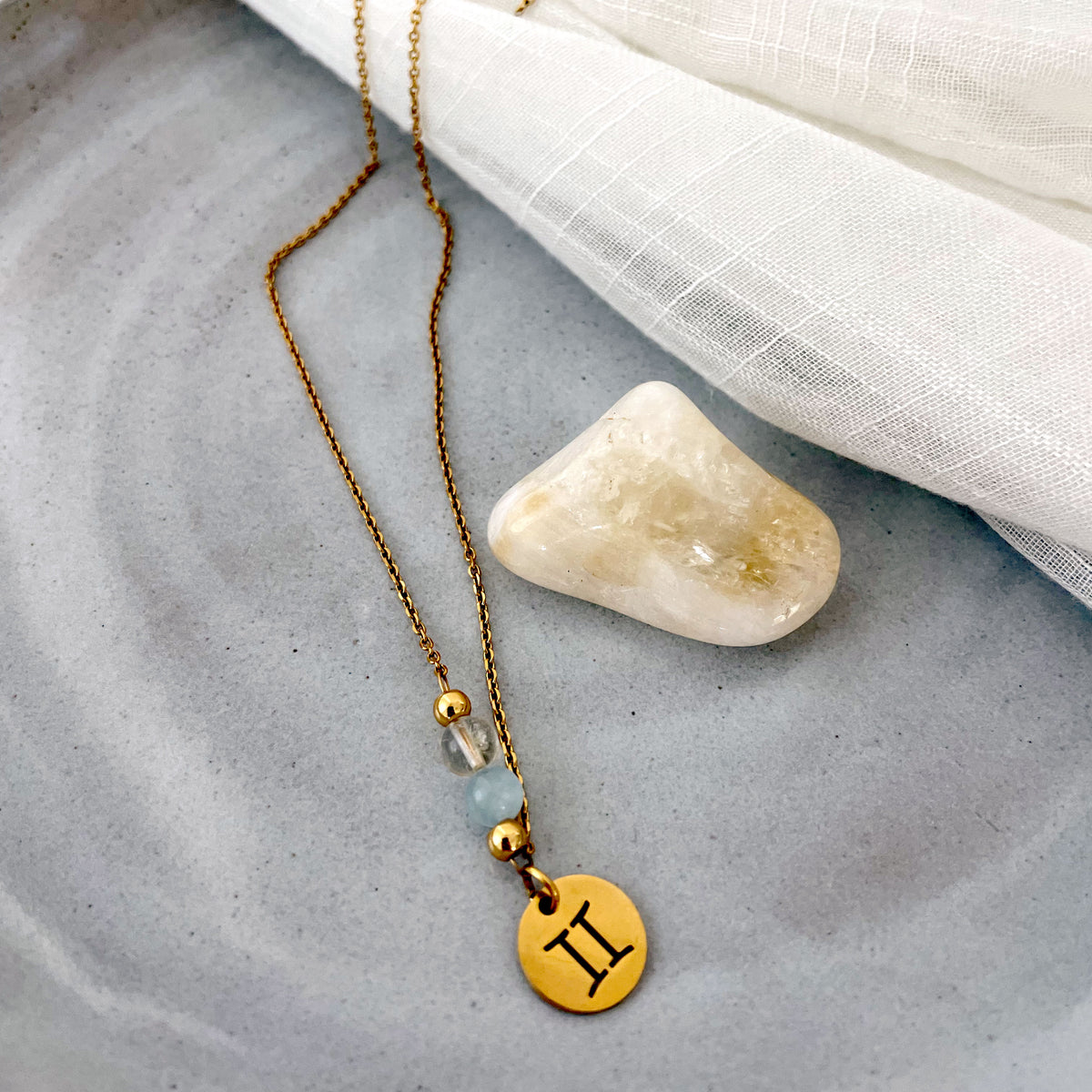 A close-up of a fine golden necklace with a gemini pendant, a Citrine and a Aquamarine bead on a ceramic plate with a Citrine gemstone.