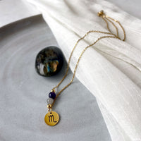 A fine golden necklace with a Labradorite and an Amethyst bead and a Scorpio Zodiac pendant laying on a light fabric. Next to the necklace is a Labradorite gemstone.