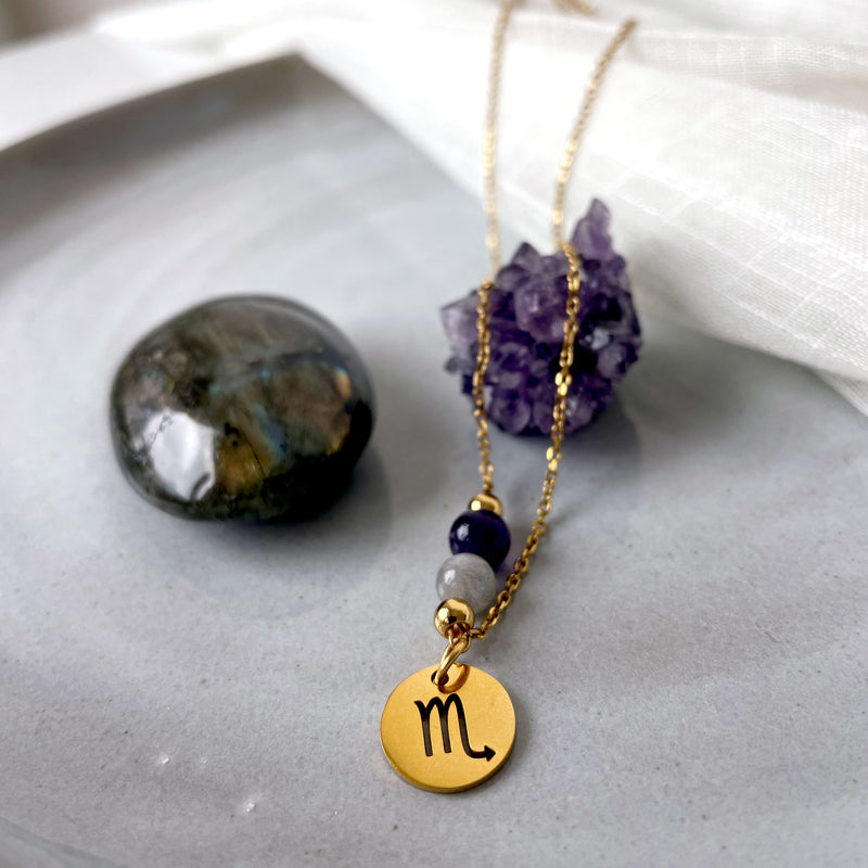 A close-up of a fine golden necklace with a Labradorite and an Amethyst bead and a Scorpio Zodiac pendant laying on a light fabric. The necklace is draped on an Amethyst druse. Next to the necklace is a Labradorite gemstone.