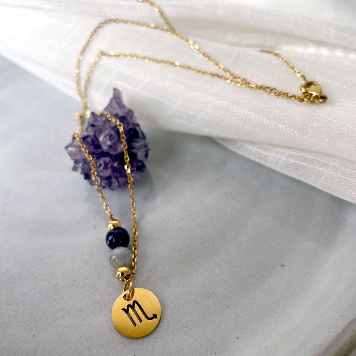 A close-up of a fine golden necklace with a Labradorite and an Amethyst bead and a Scorpio Zodiac pendant laying on a light fabric. The necklace is draped on an Amethyst druse.