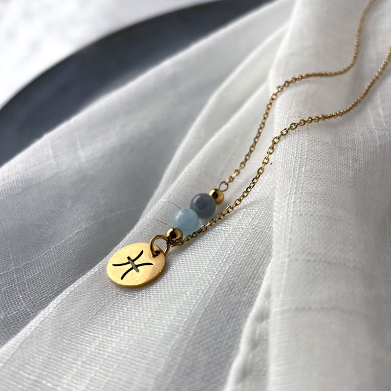 A close-up of a fine golden necklace with a Labradorite and an Aquamarine bead and a Pisces Zodiac pendant laying on a light fabric.