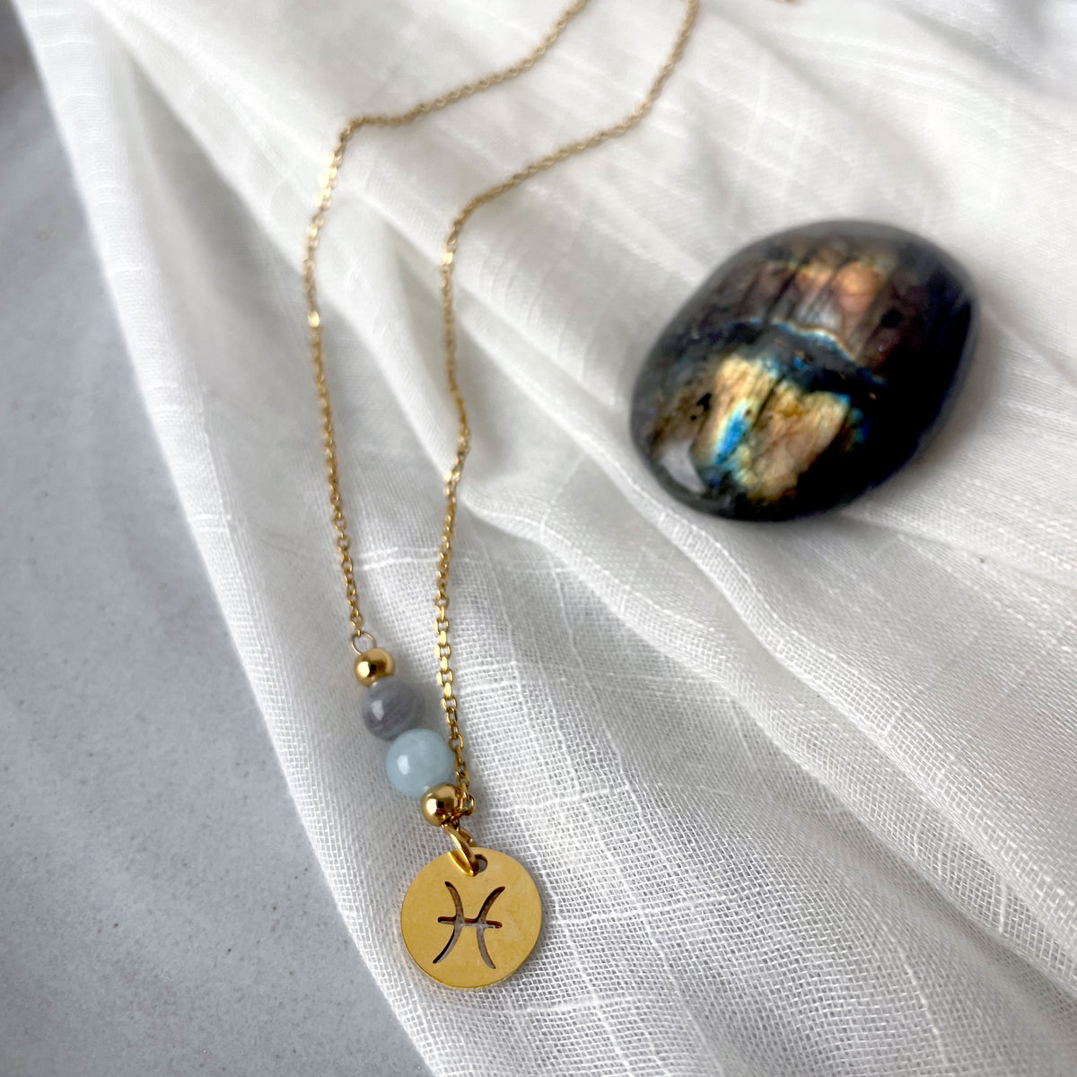 A fine golden necklace with a Labradorite and an Aquamarine bead and a Pisces Zodiac pendant laying on a light fabric. Next to the necklace is a Labradorite.