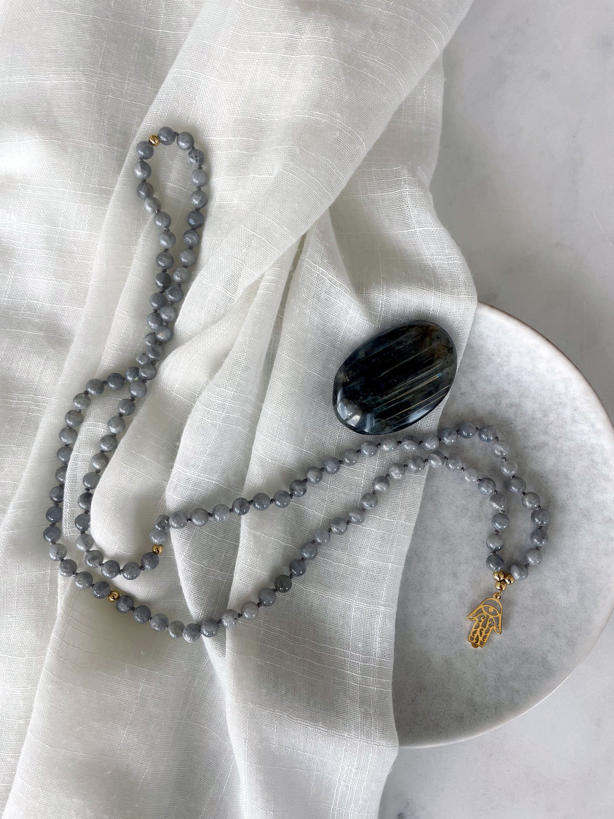 A Mala made of Labradorite gemstones and a gold plated Hamsa Hand as a pendant next to a Labradorite gemstone on a grey ceramic plate with a light linen fabric.