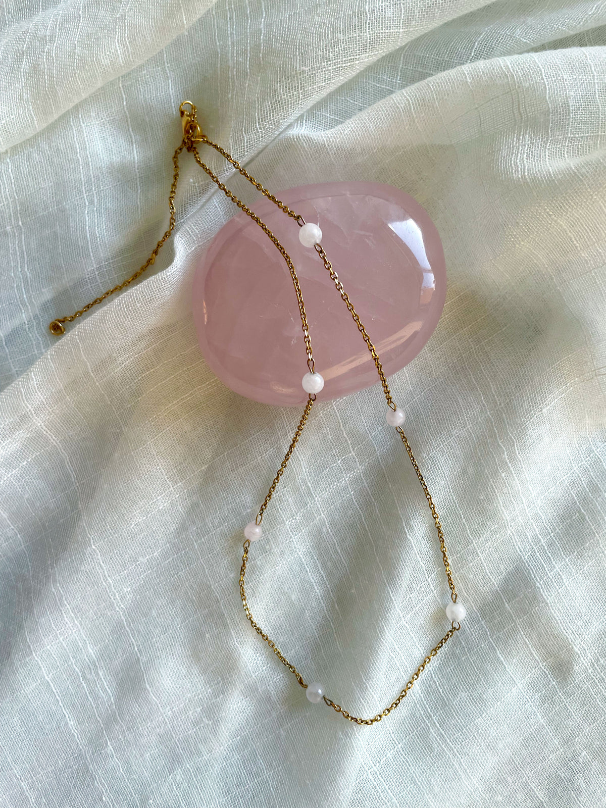 A fine golden necklace with 6 4mm Rose Quartz beads on a light white fabric and a Rose Quartz gemstone.