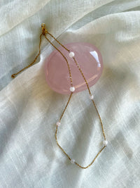 A fine golden necklace with 6 4mm Rose Quartz beads on a light white fabric and a Rose Quartz gemstone.