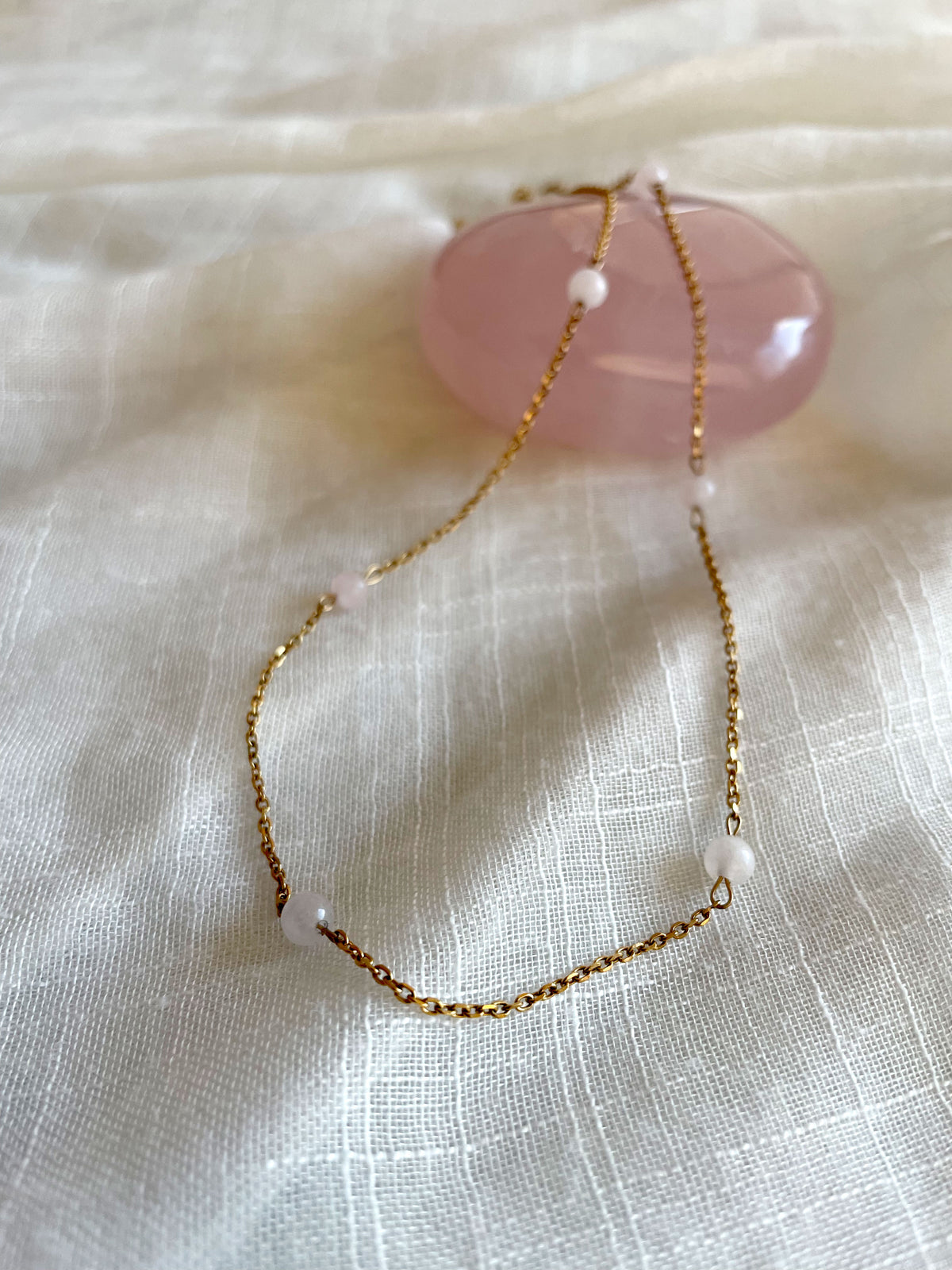 A fine golden necklace with 6 4mm Rose Quartz beads on a light white fabric and a Rose Quartz gemstone.