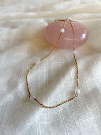 A fine golden necklace with 6 4mm Rose Quartz beads on a light white fabric and a Rose Quartz gemstone.