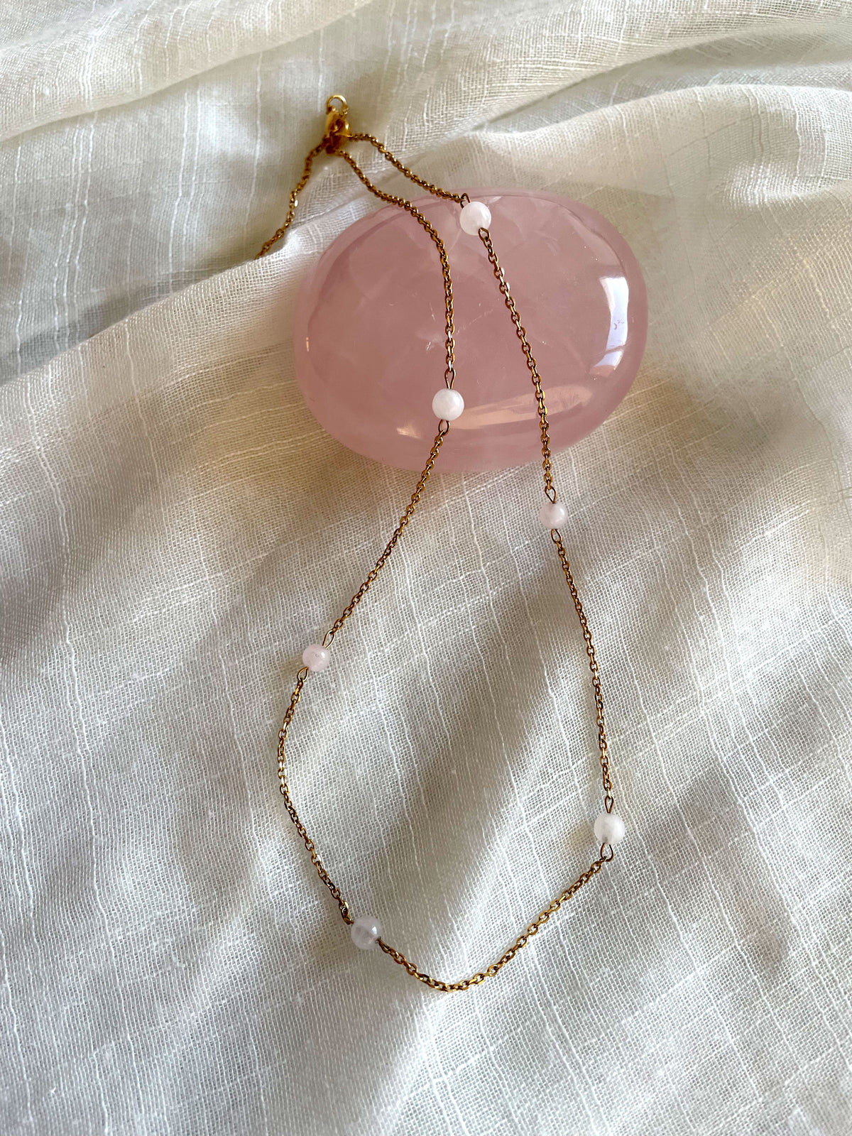 A fine golden necklace with 6 4mm Rose Quartz beads on a light white fabric and a Rose Quartz gemstone.