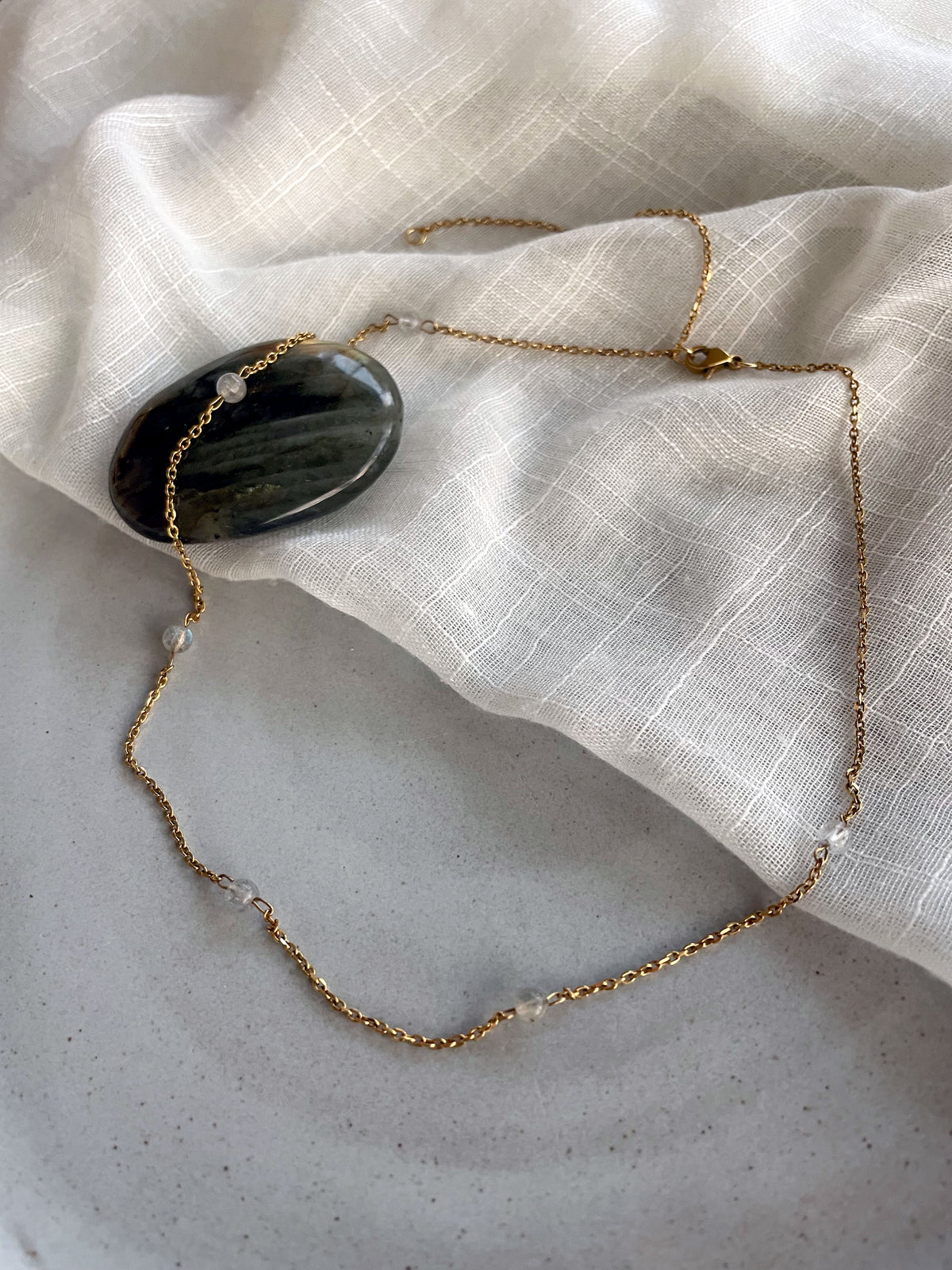 A fine golden necklace with 4mm Labradorite beads on a ceramic with a white fabric with a Labradorite gemstone.