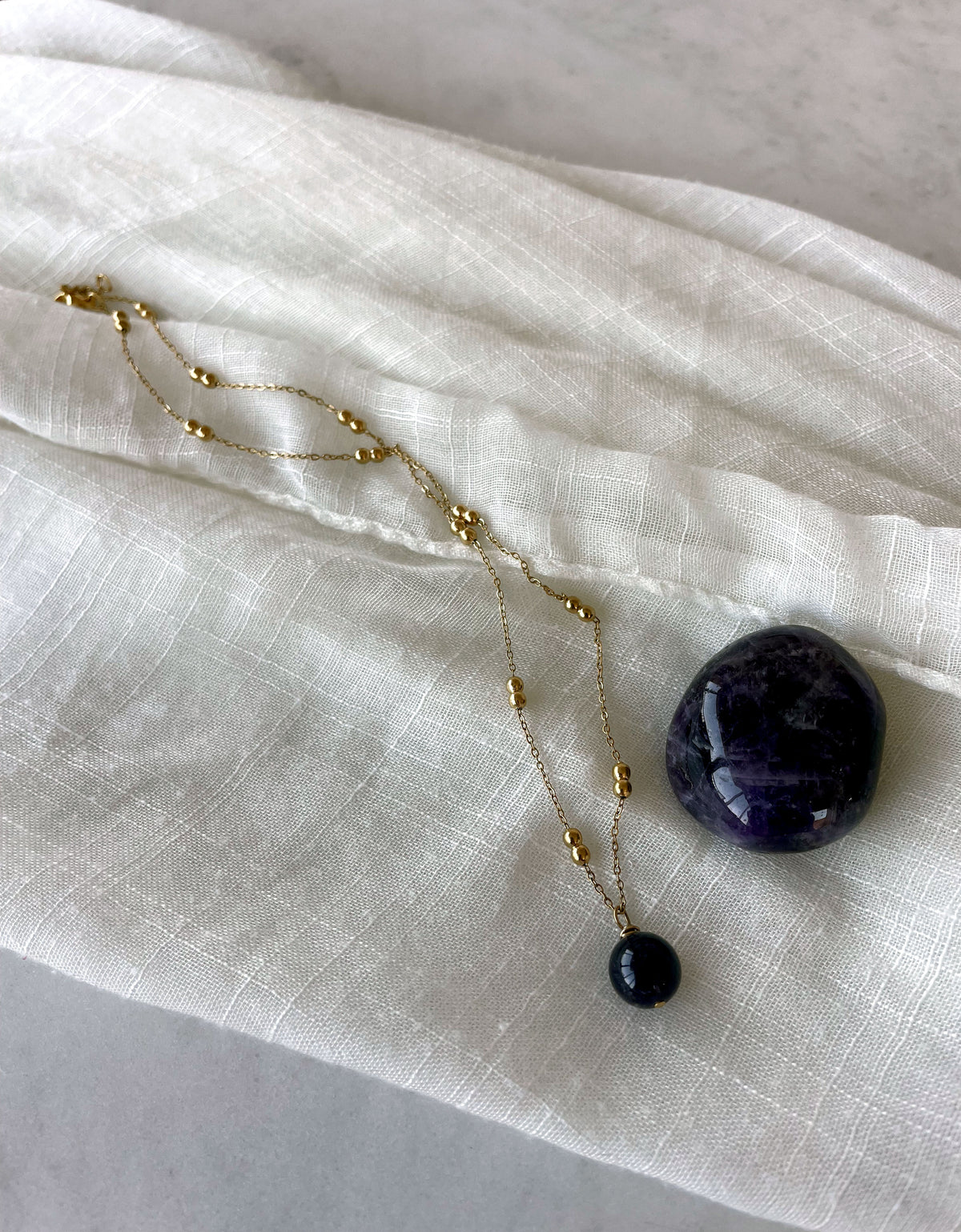 A gold plated necklace with a 10mm Amethyst bead on a delicate chain with 3mm Ball and 1mm Curb Chain on a light linen fabric.