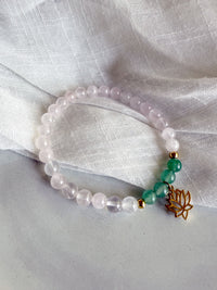 “I Am Love” heart chakra bracelet handcrafted by Moksha Jewelry with rose quartz and green aventurine beads, gold-plated accents, and a lotus charm, displayed on a soft gray surface with textured white fabric—symbolizing self-love, compassion, and emotional balance.