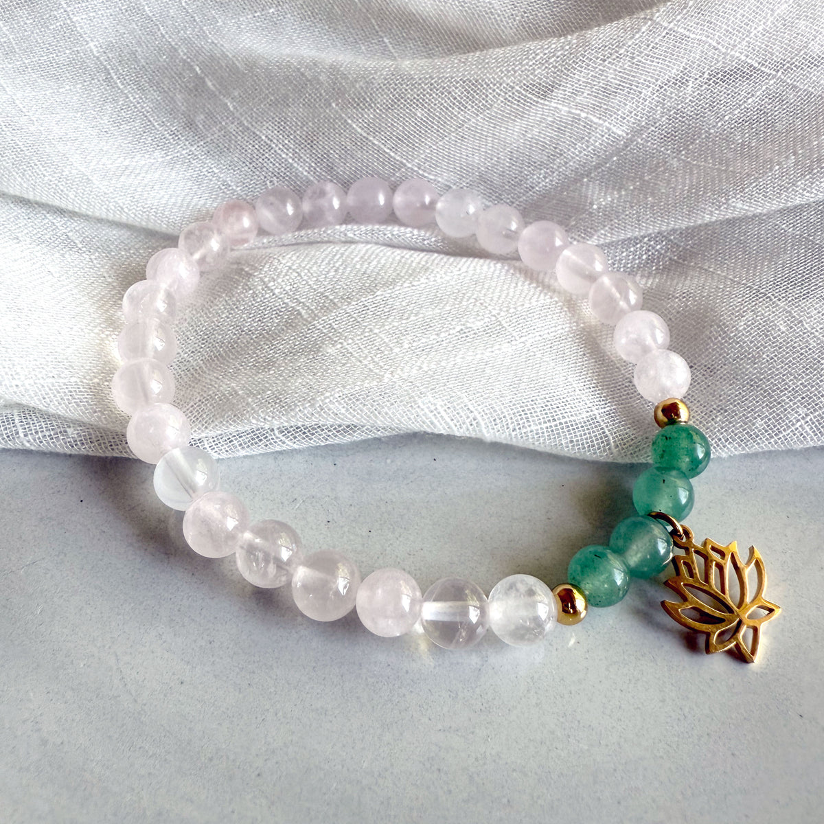 I Am Love” heart chakra bracelet by Moksha Jewelry, handcrafted with rose quartz and green aventurine beads, accented with gold details and a lotus charm, styled on a soft gray surface with a textured white fabric—designed to inspire love, balance, and emotional healing.